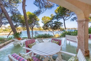 An outdoor seating area with a table and chairs, surrounded by trees and overlooking a body of water. - Paraiso sobre el Mar (Urbanización de la Torre, Porto Petro, Santanyí)