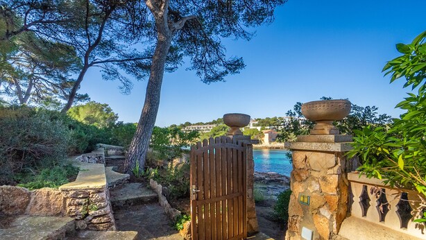 A stone gate leading to a pathway with a view of a body of water and buildings in the background. - Paraiso sobre el Mar (Urbanización de la Torre, Porto Petro, Santanyí)