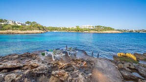 A coastal view with a rocky shoreline, clear blue water, and buildings in the background. - Paraiso sobre el Mar (Urbanización de la Torre, Porto Petro, Santanyí)