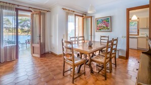 A dining area with a wooden table and chairs, a painting on the wall, and a view of the outdoors. - Paraiso sobre el Mar (Urbanización de la Torre, Porto Petro, Santanyí)