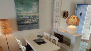 A dining area with a white table and chairs, a painting on the wall, a floor lamp, and a kitchen area with a white cabinet and a hanging light fixture. - La Paloma Blanca-WELCOMEPLAYABLANCA (Playa Blanca)