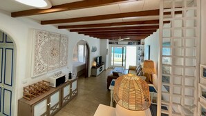 A spacious living area with a high ceiling, wooden beams, a large decorative wall piece, a dining table with chairs, and a view through a glass door to the outside. - La Paloma Blanca-WELCOMEPLAYABLANCA (Playa Blanca)