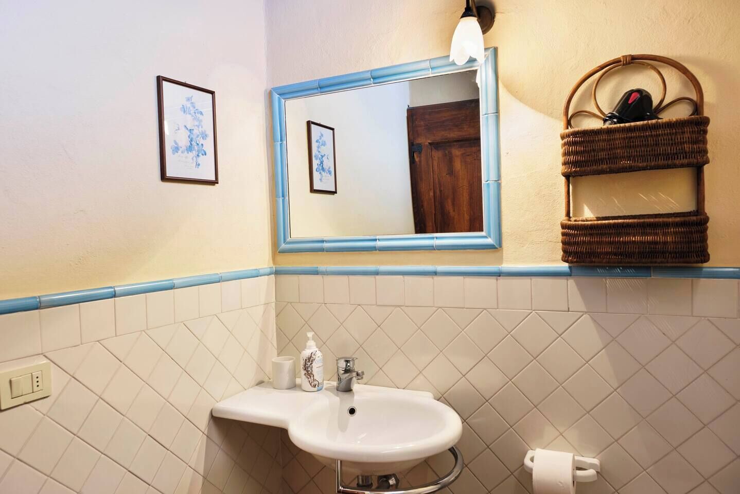 A bathroom with a white sink, a mirror, a framed picture, and a wicker basket with toiletries.