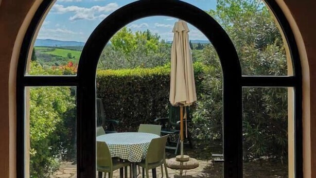 A view through a large arched window of a stone patio with a table and chairs, an umbrella, and lush greenery. - Glicini 1, holiday home with pool 30' from Florence (Barberino Tavarnelle)