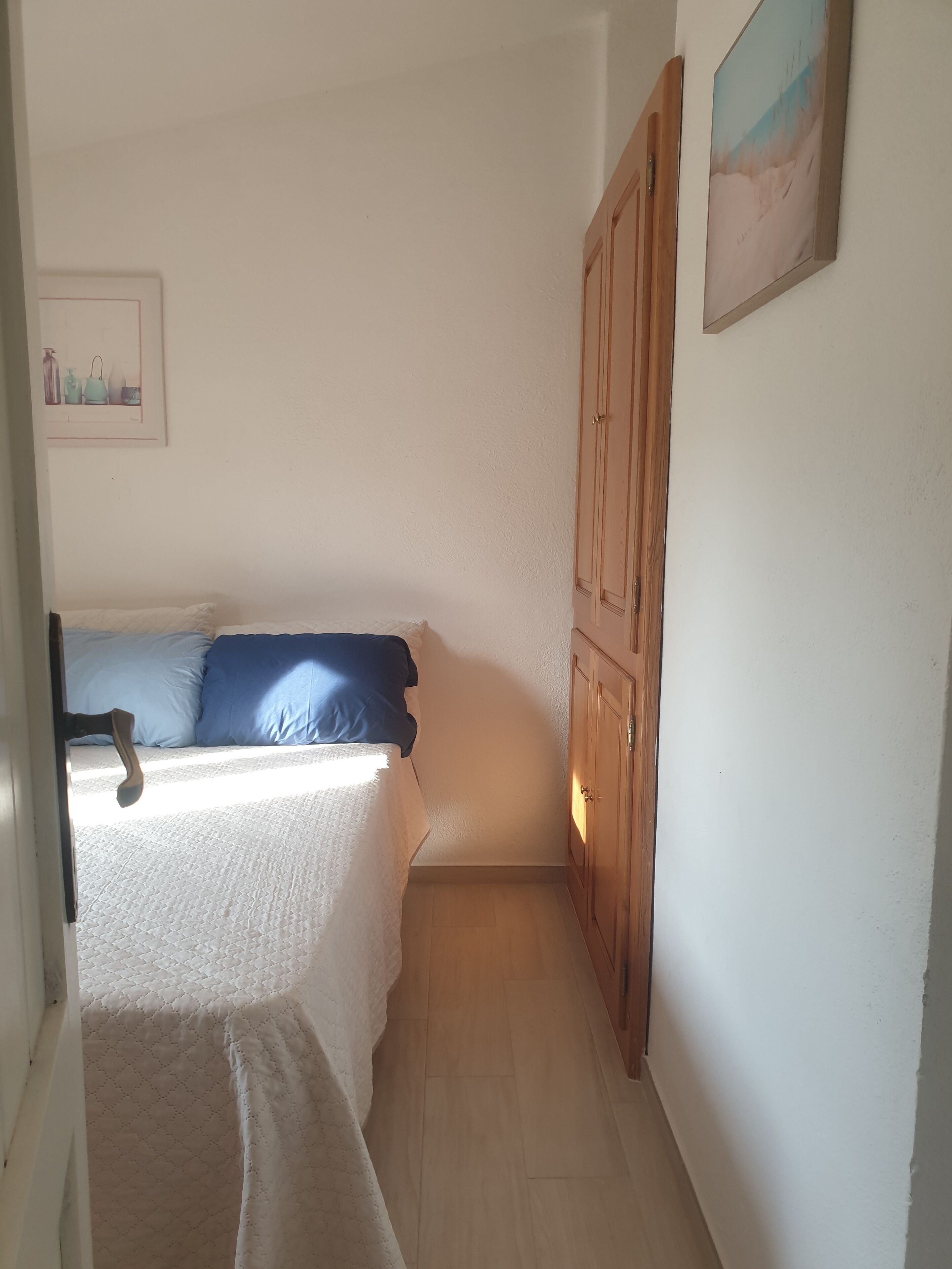 A narrow hallway leading to a bedroom with a bed, a wooden wardrobe, and a framed picture on the wall.