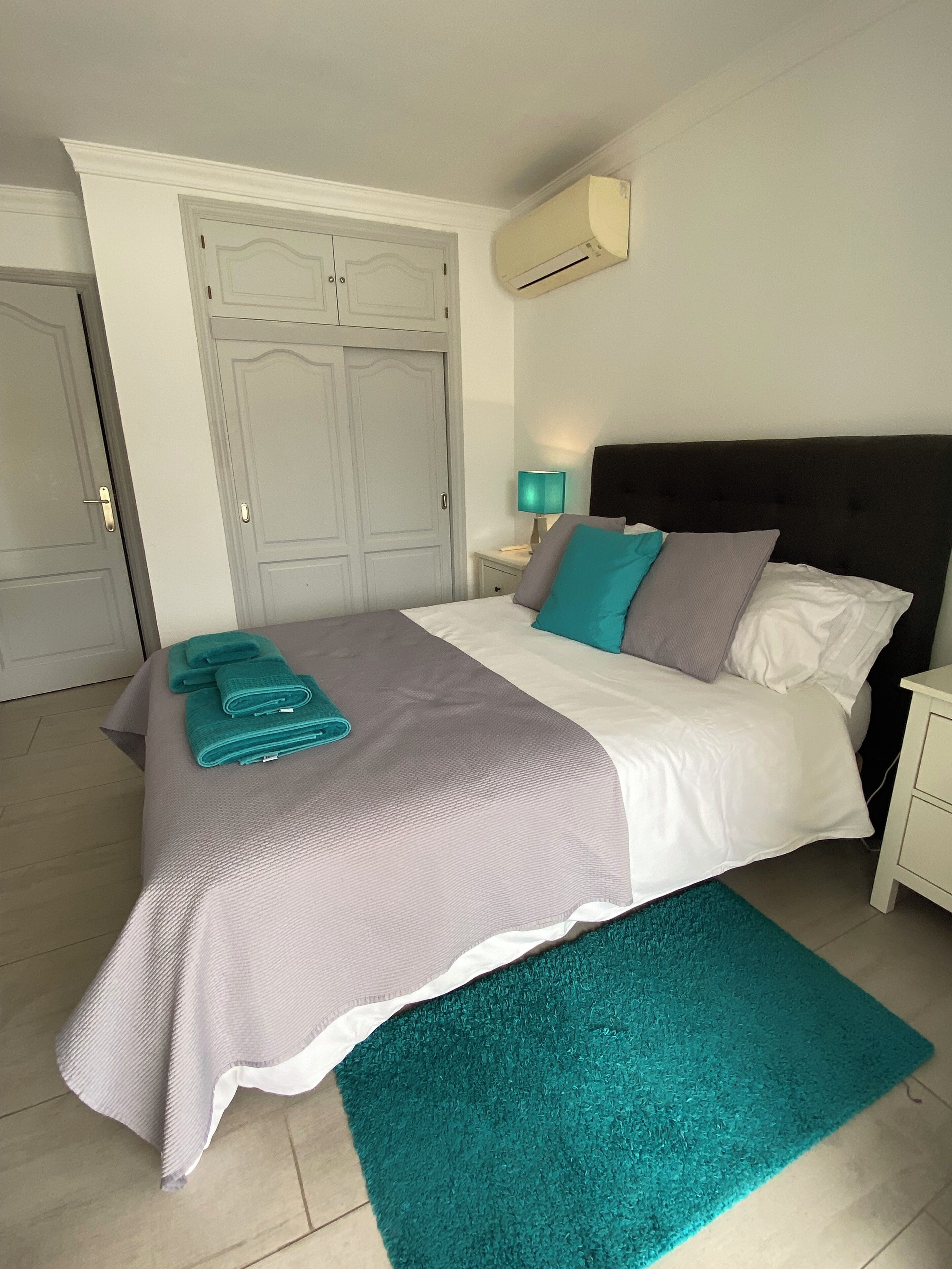 A neatly made bed with a teal throw blanket and pillows, a teal rug, a bedside lamp, and an air conditioning unit on the wall.