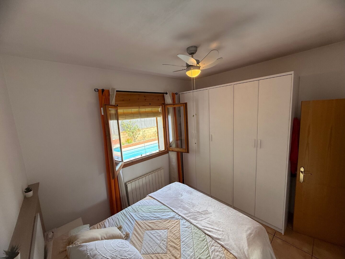 Une chambre à coucher comprenant un lit, un ventilateur de plafond, une fenêtre avec des rideaux, une armoire et un radiateur.