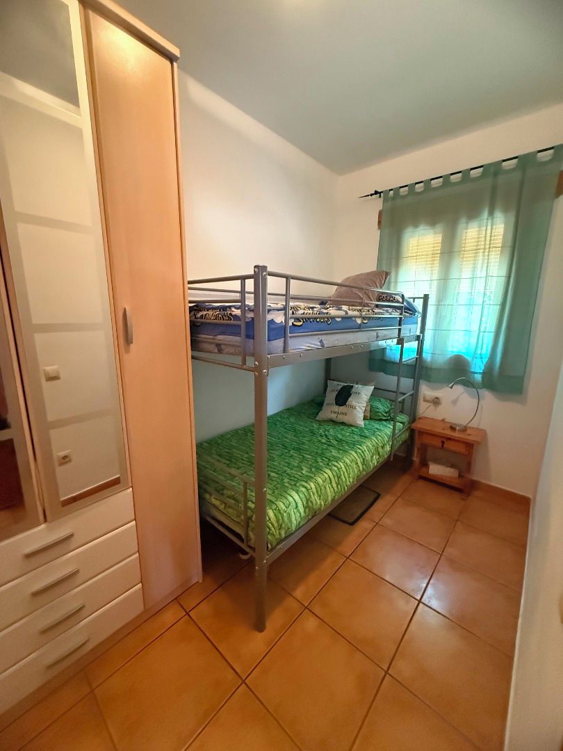 A room with a bunk bed, a wooden wardrobe, a small bedside table, and a window with curtains.