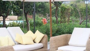 Eine Lounge-Ecke mit Korbmöbeln und weißen Kissen, einem großen Sonnenschirm sowie Blick auf Grünflächen und einen Pool im Hintergrund.