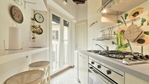 A kitchen with a gas stove, oven, and a sink. There are two wicker stools and a white shelf with decorative items. - Wine Apartments Florence Trebbiano (Florence)