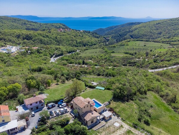An aerial view of a rural area with a large house featuring a swimming pool, surrounded by lush greenery and a view of the sea.