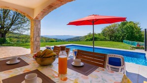 Zona de menjador a l'aire lliure amb vista a la piscina, un parasol vermell i una taula preparada amb fruita i begudes.