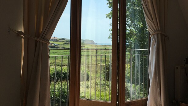 A room with a large glass door leading to a balcony with a view of greenery and distant hills. - House With Sea Views And Sunny Aspect (Thurlestone)