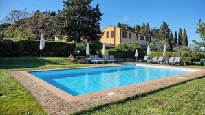 A rectangular swimming pool with a stone border, surrounded by a grassy area and a row of sun loungers with white umbrellas.