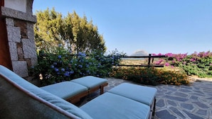 A stone patio with two cushioned seating areas, surrounded by colorful flowering plants and a view of the landscape. - Two-room apartment with sea view, swimming pool and relax (Porto San Paolo)