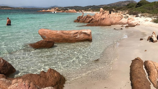 A beach with clear turquoise water, large rocks, and a distant view of mountains. - Two-room apartment with sea view, swimming pool and relax (Porto San Paolo)