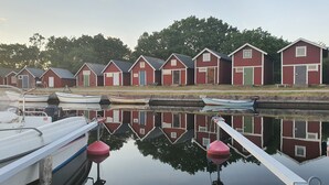 Une rangée de bâtiments en bois peints en rouge, dotés de fenêtres et de portes, se reflète dans l’eau calme. Des bateaux sont amarrés à proximité.