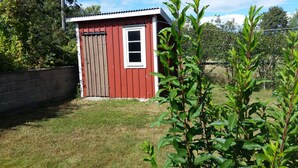 Un petit abri en bois rouge, avec une fenêtre blanche et une porte, entouré d’herbe et de plantes.