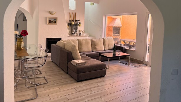 Un salon moderne avec un canapé d’angle, une table à manger en verre et des chaises. On y trouve une cheminée, une grande fenêtre donnant sur une structure en briques à l’extérieur, et une petite table sur laquelle est posé un vase de fleurs.