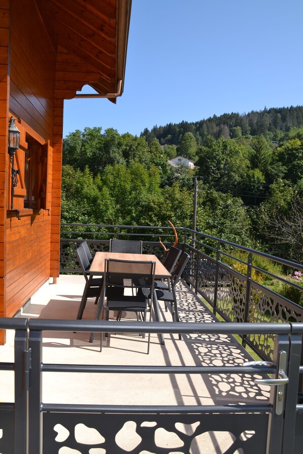 A wooden cabin with a balcony, outdoor seating, and a view of a forested hillside.