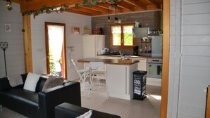 A cozy cabin interior with a living area featuring a sofa and chairs, a wooden dining table, and a kitchen with a refrigerator, stove, and cabinets.