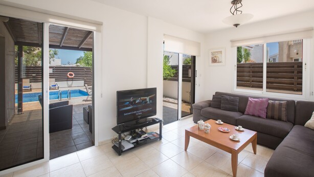 Ein Wohnzimmer mit Fernseher, Couch und Couchtisch. Durch die Glastüren ist ein Pool zu sehen.