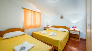 A room with two beds, each with a yellow bedspread and white pillows, a small bedside table with a lamp, and a window with an orange curtain. - ALEKS house with apartments / Aleks 2 APARTMENT (Umag)