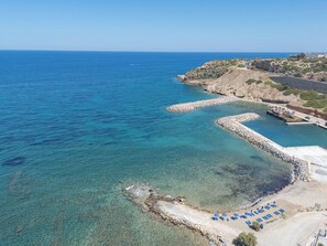 A coastal area with clear blue waters, a rocky shoreline, and a paved path leading to a beach with blue umbrellas. - La Casa di Mare, Luxury SeaFront Villa with Private Pool, Whirlpools & Sauna (Rethymno)