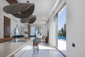 A modern kitchen with a central island, a dining area with a view of the pool, and a large sliding glass door leading to a balcony. - La Casa di Mare, Luxury SeaFront Villa with Private Pool, Whirlpools & Sauna (Rethymno)