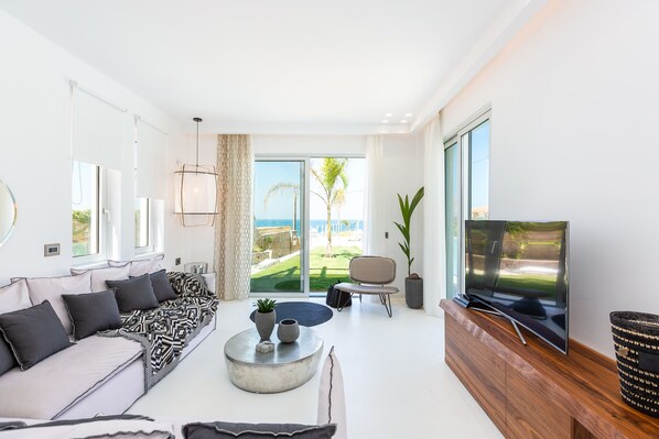 A modern living room with a sofa, coffee table, TV, and large windows overlooking a garden. - La Casa di Mare, Luxury SeaFront Villa with Private Pool, Whirlpools & Sauna (Rethymno)