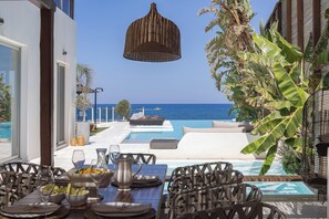 A poolside dining area with a view of the sea, outdoor furniture, and a hanging light fixture. - La Casa di Mare, Luxury SeaFront Villa with Private Pool, Whirlpools & Sauna (Rethymno)