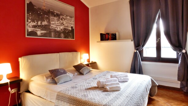 Ein Hotelzimmer mit Bett, Kissen, Decke, Nachttisch mit Lampe, Fenster mit Vorhängen, Bücherregal und einem gerahmten Bild an der Wand.