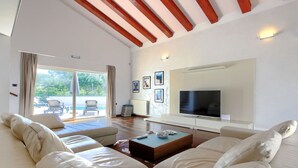 Un vaste salon comprenant un grand canapé, une table basse et un téléviseur à écran plat fixé au mur. Une porte-fenêtre coulissante donne accès à un espace extérieur aménagé pour se détendre.