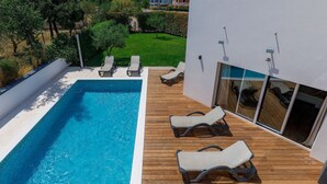Un espace piscine extérieur moderne, doté d’une terrasse en bois, de chaises longues et d’une porte vitrée donnant sur un patio abrité.