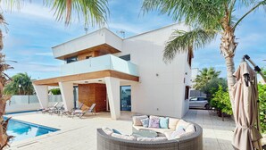 Een modern huis met twee verdiepingen, een zwembad, buitenmeubels en palmbomen.