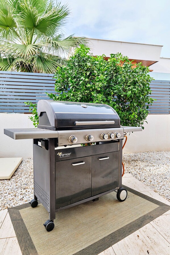 A portable gas grill with four burners, wheels, and a side shelf, placed on a patio with a pebble ground cover.