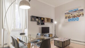 A modern living room with a glass dining table, black chairs, a floor lamp, a television, and a poster on the wall.
