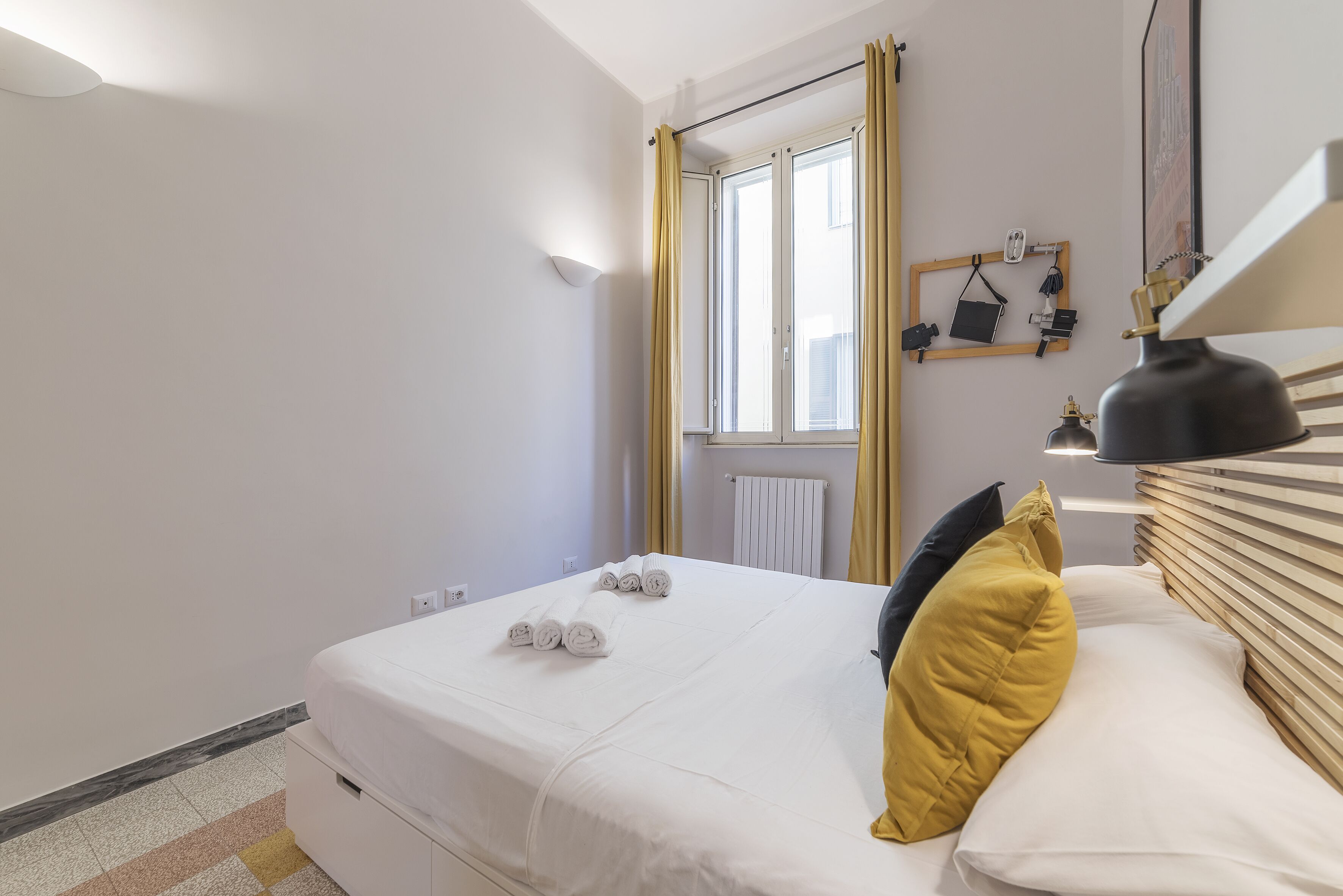 A neatly made bed with rolled towels, a bedside lamp, and a window with curtains.