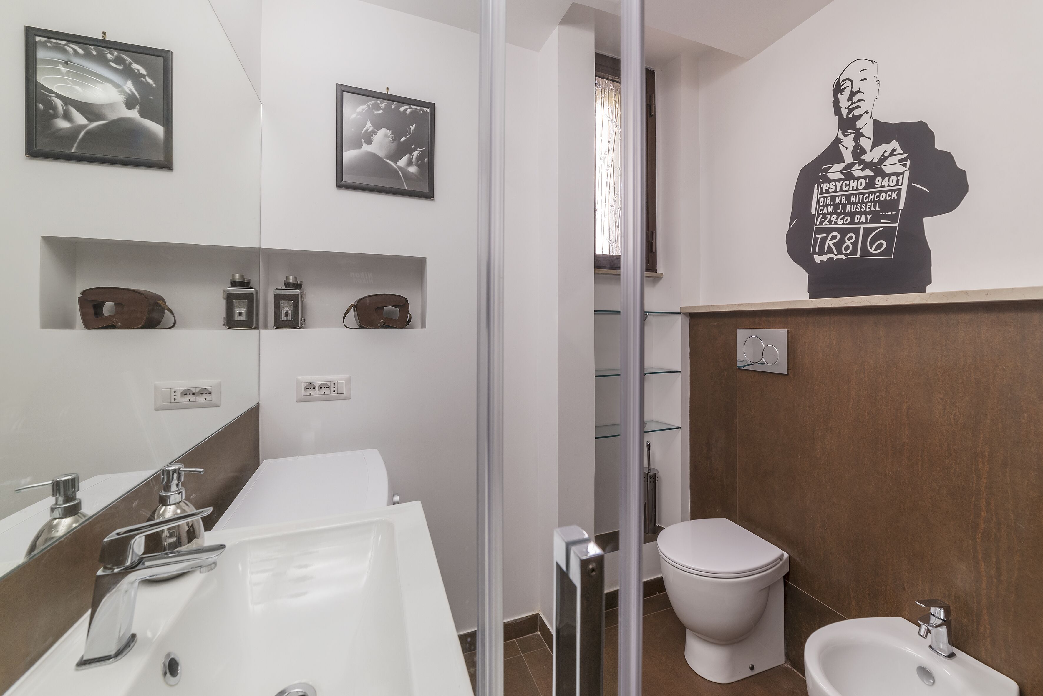 A modern bathroom with a white sink, mirror, and a bidet. There are framed pictures on the wall and a large poster.