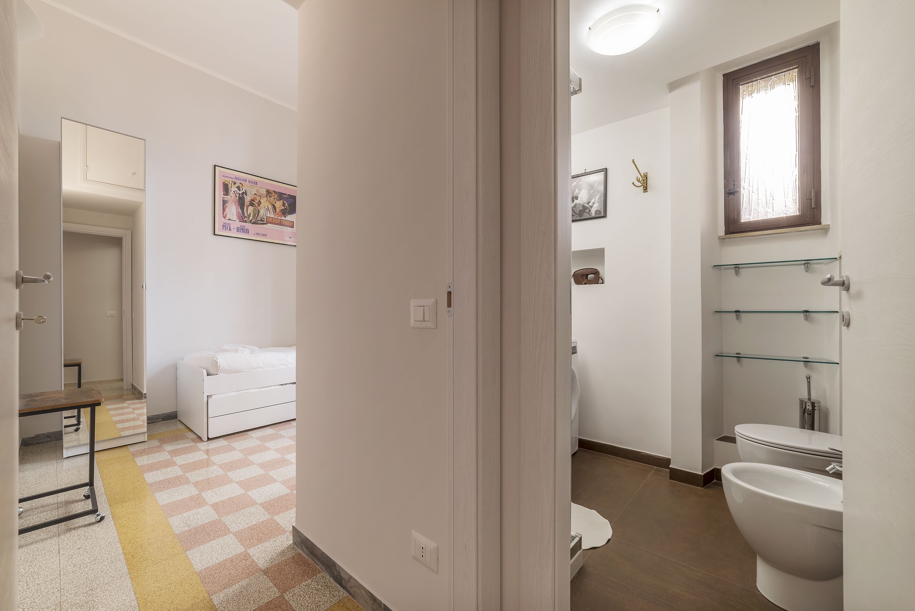 Habitación de hotel con piso a cuadros, una cama, una mesita de noche, un escritorio pequeño, una silla, una ventana con cortina, un inodoro y un lavamanos.
