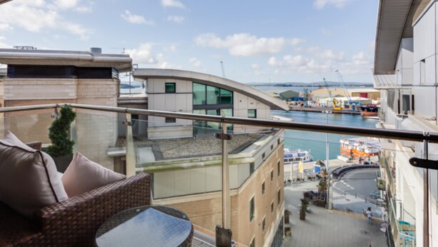 Un balcon agrémenté de meubles en osier et d’une table en verre, offrant une vue sur une marina avec des bateaux et des bâtiments.