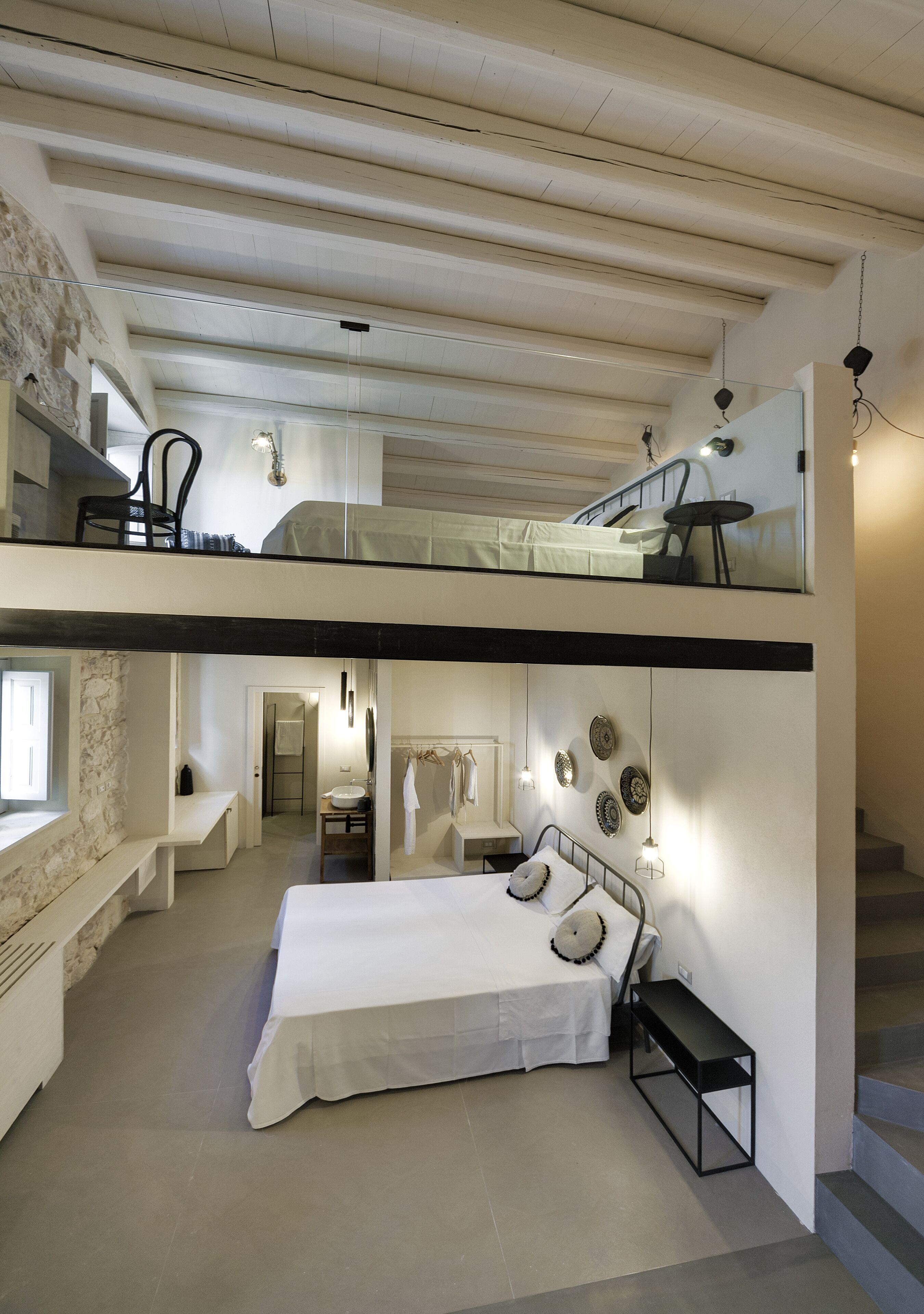 A modern, multi-level bedroom with a large bed, a small table, and a bench. There is a staircase connecting the two levels.