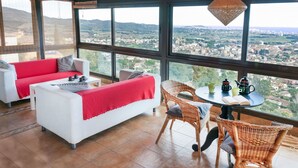 A spacious room with a large window offering a city view, a white sofa with a red throw, wicker chairs, and a small table with a teapot and cups. - Wonderful private villa with private pool, WIFI, TV, terrace, pets allowed and panoramic view (Calonge)