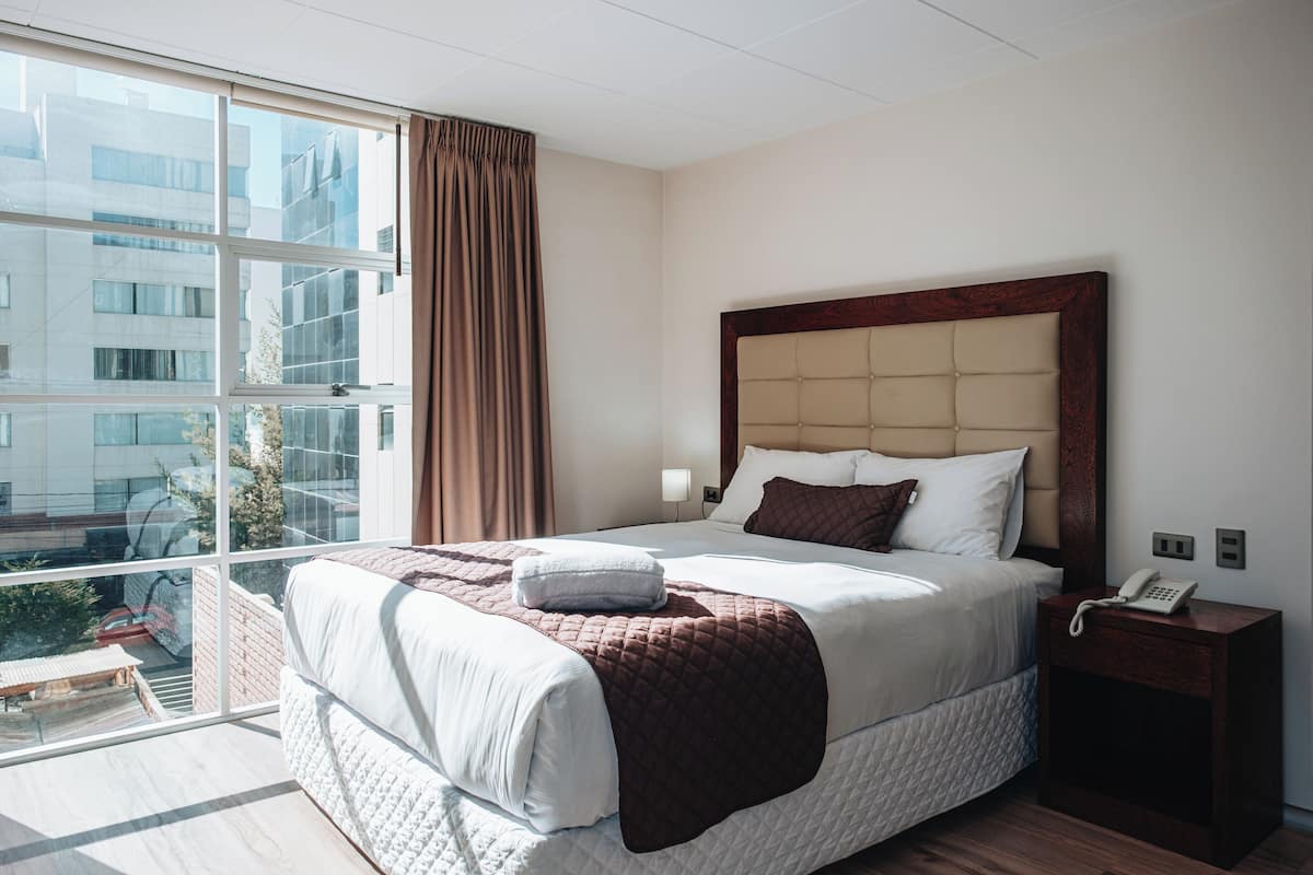 A hotel room with a large bed, a wooden headboard, a bedside table with a phone, and a view of buildings through the window.