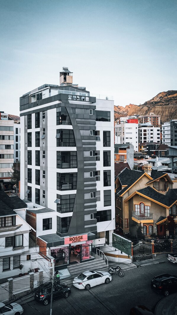 A modern multi-story building with a prominent curved facade, a visible coffee shop on the ground floor, and a mountainous backdrop. - Fratelli Aparthotel (La Paz)