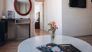 A modern hotel room with a round table, a vase of flowers, a magazine, a TV on the wall, and a wooden desk with a mirror and some items on it. - Fratelli Aparthotel (La Paz)