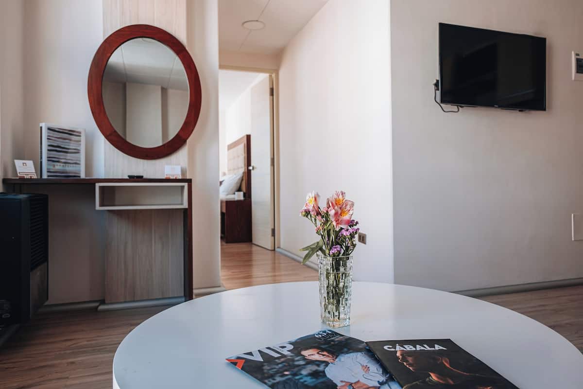 A modern hotel room with a round table, a vase of flowers, a magazine, a TV on the wall, and a wooden desk with a mirror and some items on it.