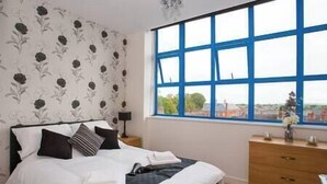 A bedroom with a large window, a bed with white and dark bedding, a wooden dresser, and a nightstand with a lamp. - Comfortable Swindon 1 bedroom Town Centre Apartment, FREE Parking (Swindon)