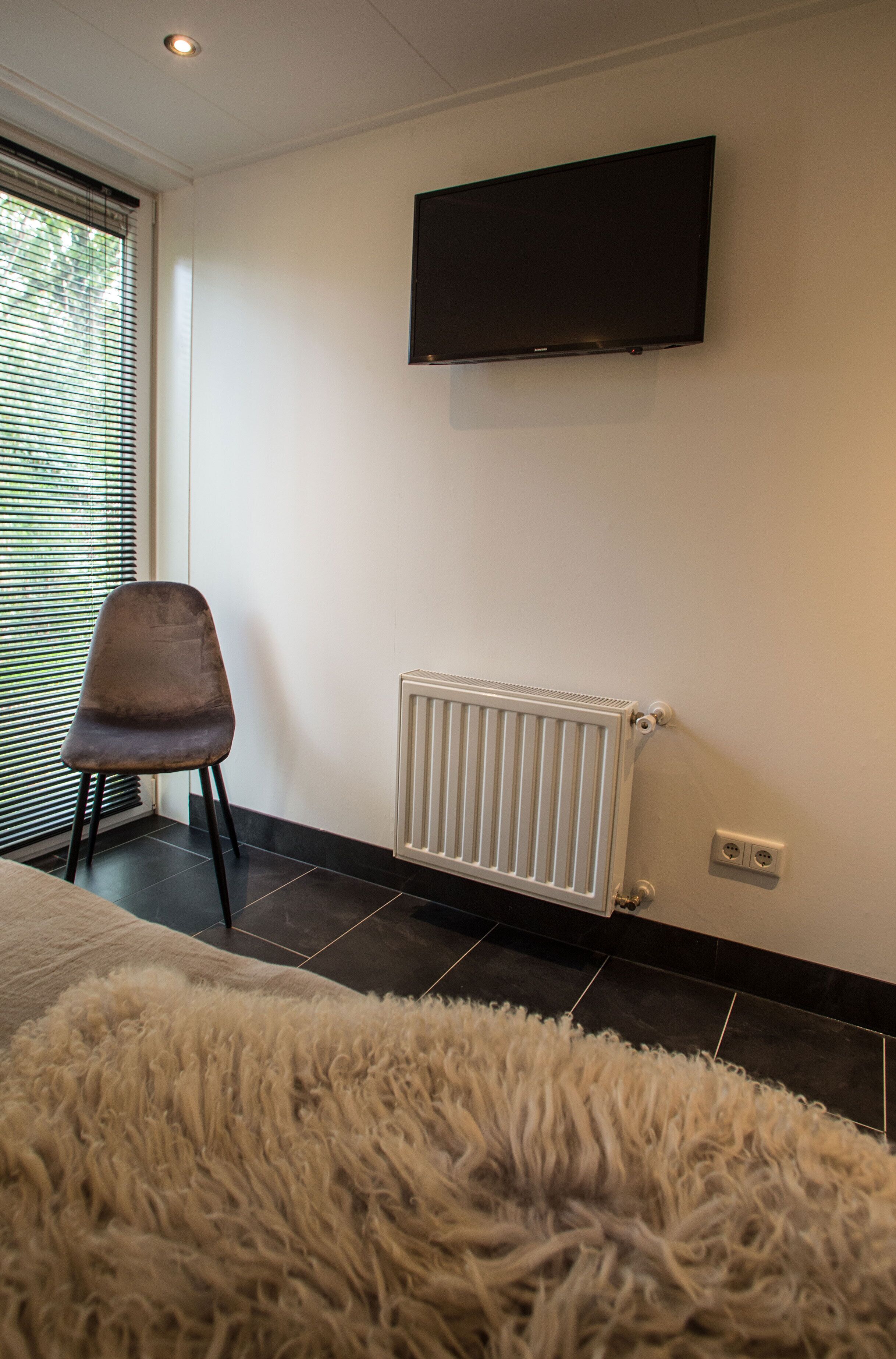 Quarto moderno com televisão de tela plana na parede, um radiador abaixo dela, uma cadeira e um tapete macio no chão.