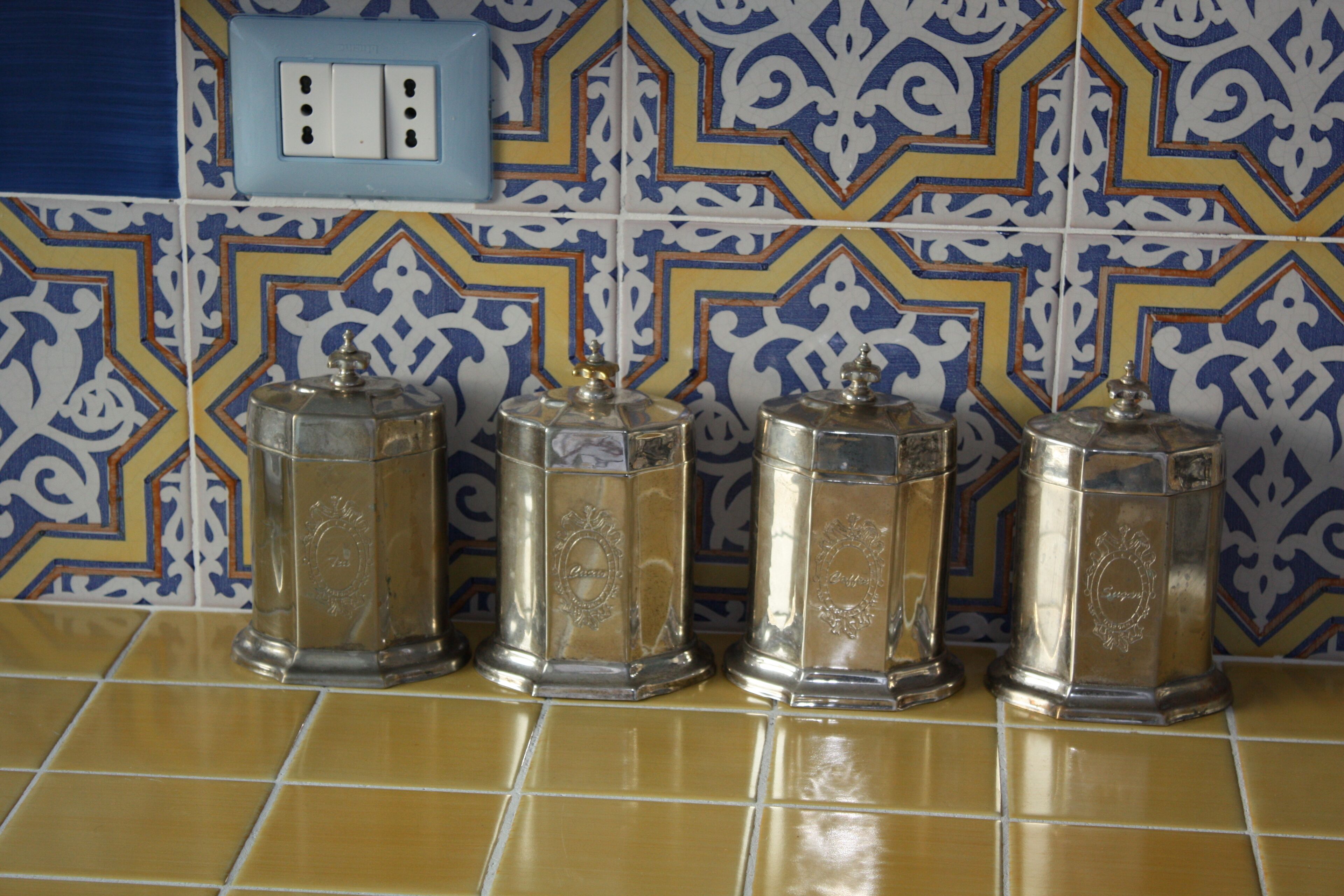 A tiled kitchen backsplash with a geometric pattern and four metallic containers on a tiled countertop.